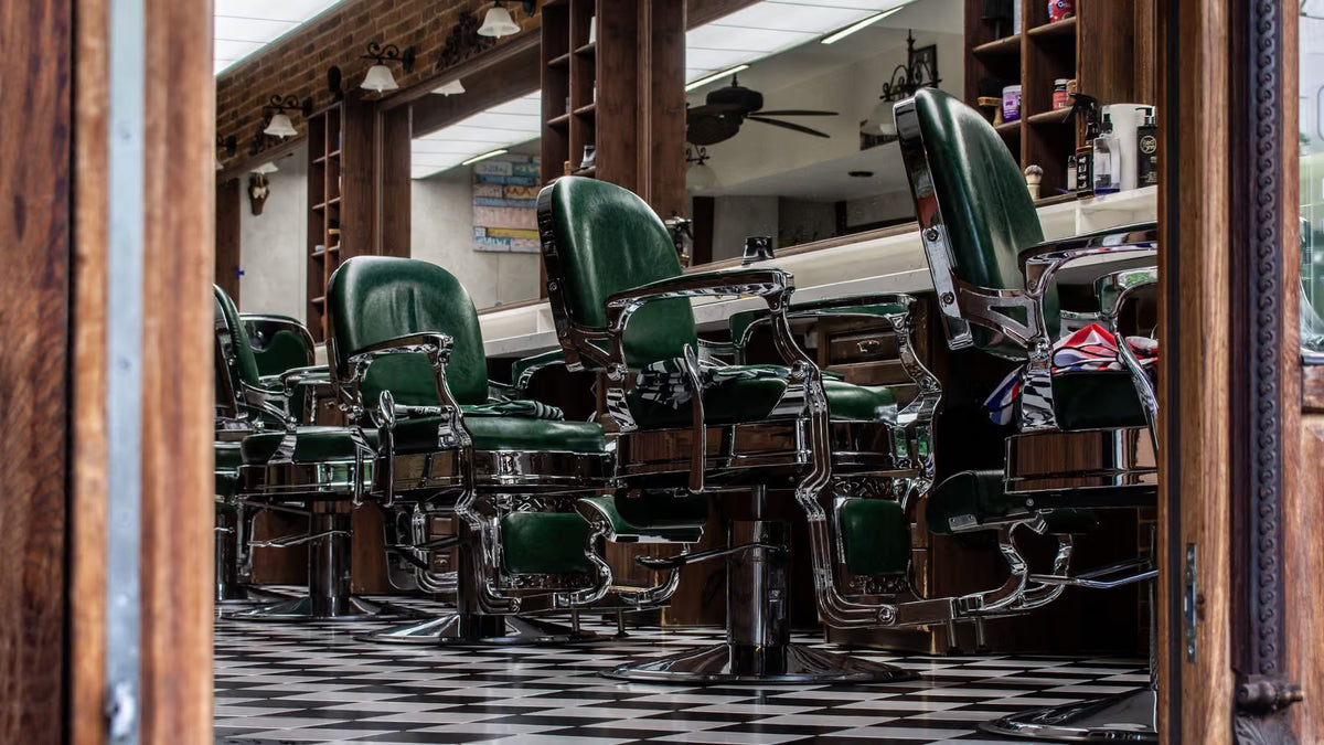 A row of classic green leather barber chairs with chrome bases arranged in a vintage barbershop, viewed through a wooden doorway. The shop has a black-and-white checkered floor, wooden shelving, and mirrors reflecting the interior.