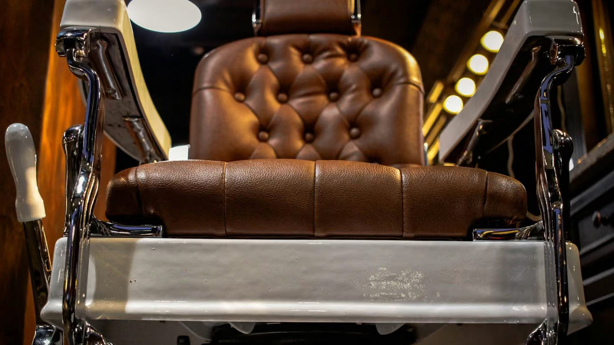 A luxury barbershop barber chair with brown tufted leather upholstery and polished chrome metal arms, shown in a dimly lit barbershop interior.